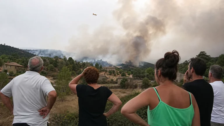 Un grupo de vecinos de un pueblo rural, con rostros de preocupación y frustración, observan a lo lejos una columna de humo elevándose sobre un bosque quemado. En el cielo, un pequeño hidroavión apenas visible, simbolizando la insuficiencia de medios. La escena transmite desolación y abandono.