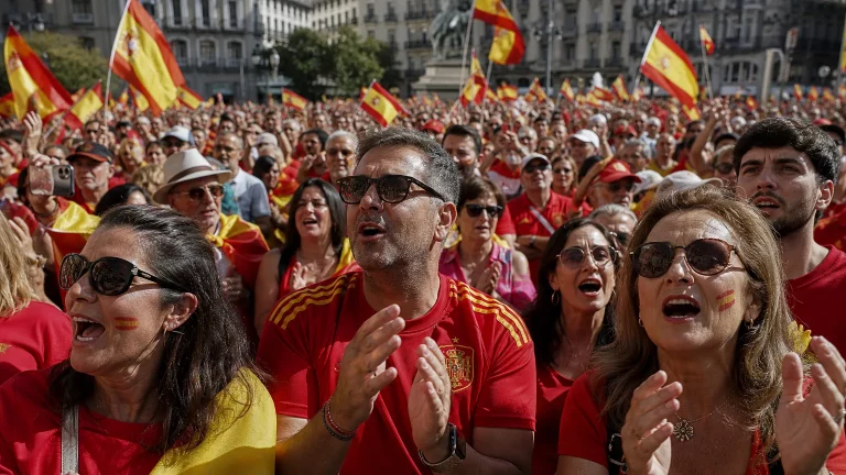 Multitud de aficionados españoles celebrando una victoria de fútbol, vestidos con camisetas rojas, ondeando banderas de España, con rostros de alegría y unidad.