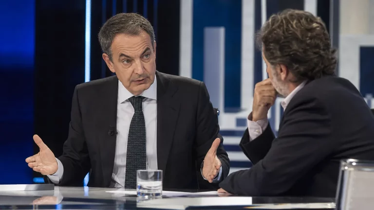 Composición editorial sobria: José Luis Rodríguez Zapatero en un plató de televisión, con gesto evasivo, mientras un periodista le observa con una expresión de interrogación. Al fondo, una silueta difusa de Pedro Sánchez.
