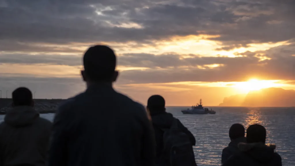 Siluetas de personas observando la costa de Ceuta al atardecer, con una patrullera de fondo en el mar, evocando una situación fronteriza delicada.