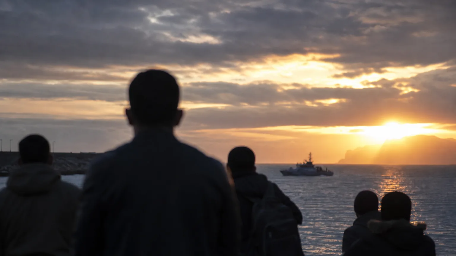 Siluetas de personas observando la costa de Ceuta al atardecer, con una patrullera de fondo en el mar, evocando una situación fronteriza delicada.