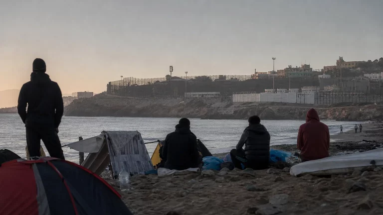 Composición editorial sobria de personas en silueta en una playa, simbolizando la situación de los inmigrantes en Ceuta, con una valla fronteriza al fondo y el mar en primer plano.
