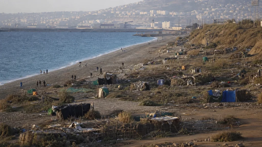Vista aérea o panorámica de la costa de Ceuta, mostrando una zona de asentamientos precarios con pequeñas cabañas construidas con ramas y materiales de desecho, y siluetas de personas moviéndose en la distancia.