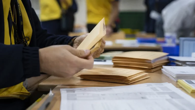 Manos anónimas de un funcionario de Correos manipulando sobres de voto por correo sobre una mesa, con documentos electorales desenfocados en el fondo.