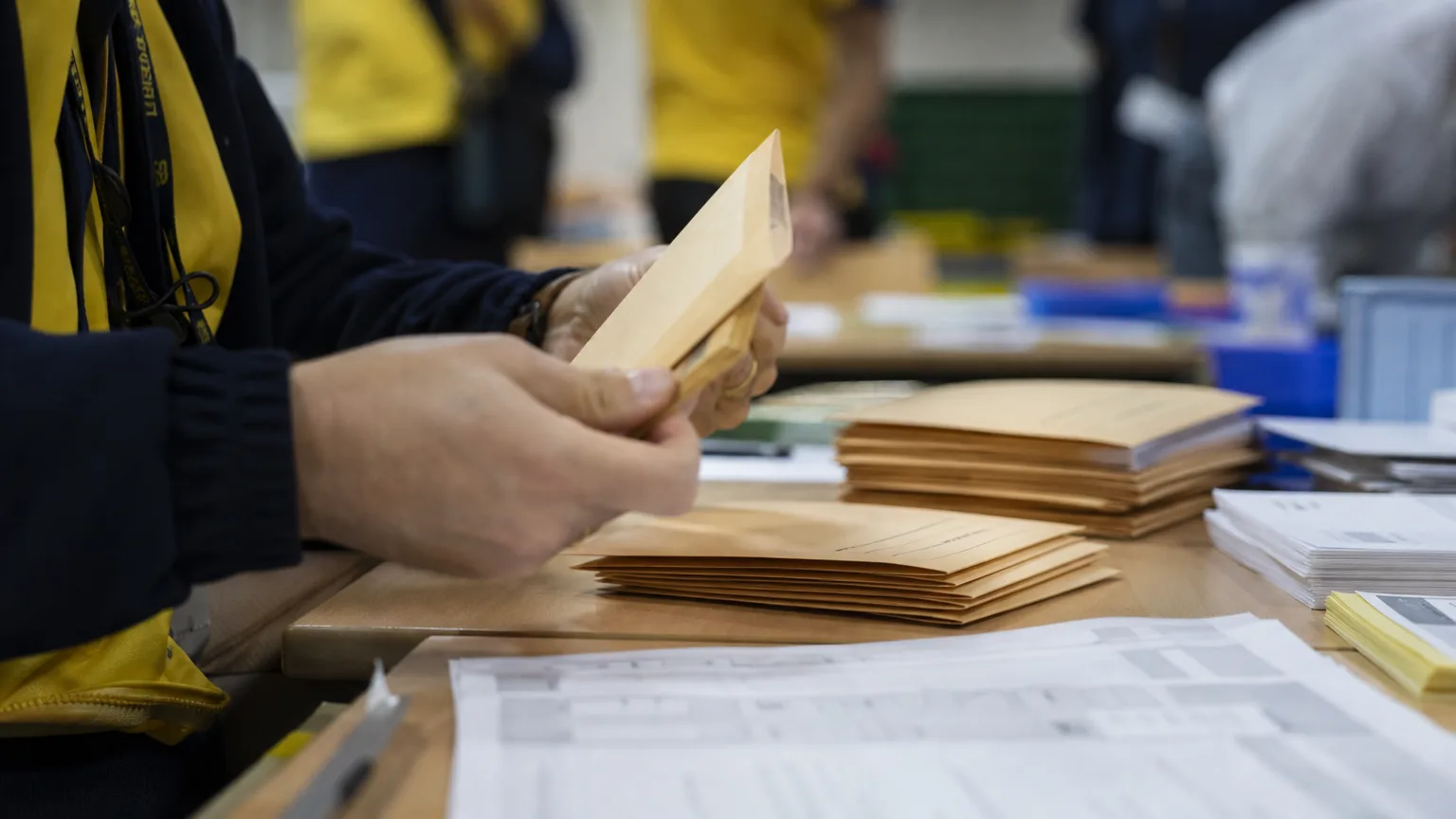 Manos anónimas de un funcionario de Correos manipulando sobres de voto por correo sobre una mesa, con documentos electorales desenfocados en el fondo.