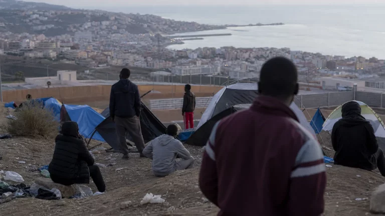 Vista aérea parcial de un campamento improvisado en una zona costera de Ceuta, con siluetas de personas de espaldas y algunas tiendas de campaña, mostrando un ambiente de precariedad y abandono, con la ciudad al fondo.