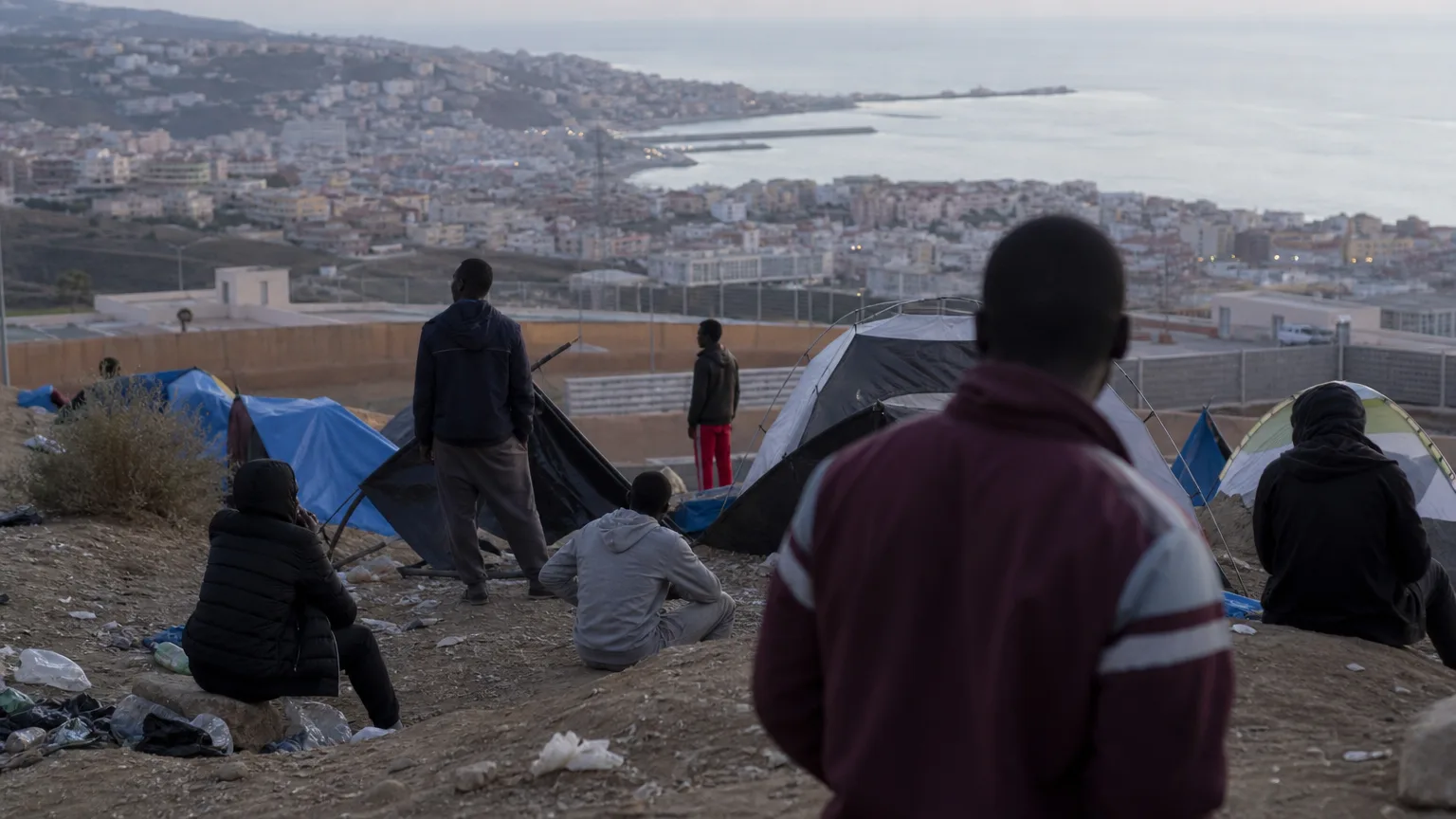 Vista aérea parcial de un campamento improvisado en una zona costera de Ceuta, con siluetas de personas de espaldas y algunas tiendas de campaña, mostrando un ambiente de precariedad y abandono, con la ciudad al fondo.