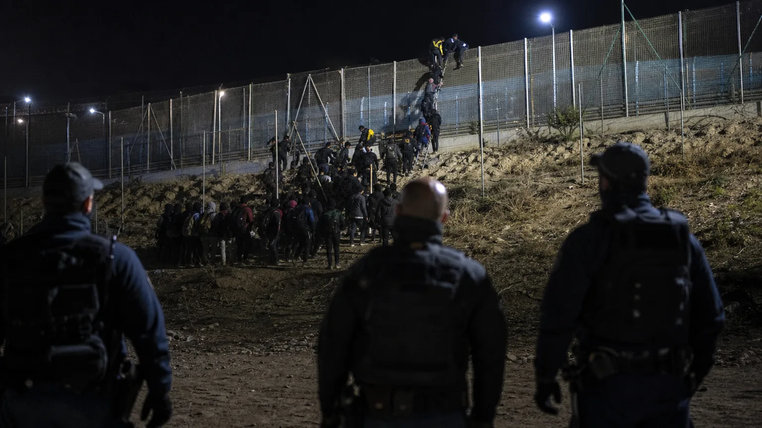 Siluetas de personas cruzando una valla en una frontera al amanecer, con una ciudad a lo lejos, representando la entrada masiva de migrantes en Ceuta. Se aprecian agentes de seguridad de espaldas en primer plano, observando la situación.