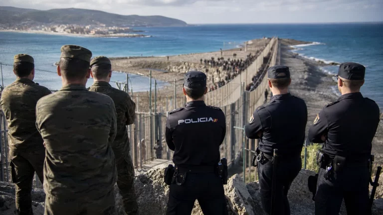Soldados y agentes de la Policía Nacional con uniformes de servicio, observando con preocupación y semblante serio el horizonte de Ceuta, con la valla fronteriza al fondo. Algunos muestran gestos de frustración o impotencia, representando la indignación por las órdenes de no intervención.