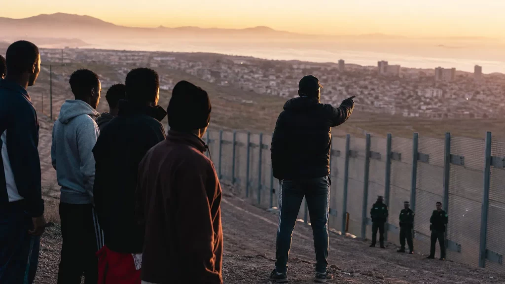 Composición editorial sobria: siluetas de personas junto a una valla fronteriza, mientras una figura en perfil parcial, que sugiere una autoridad marroquí, parece observar. En el fondo, una bandera española ondea, y figuras difuminadas de la Guardia Civil se ven pasivas.