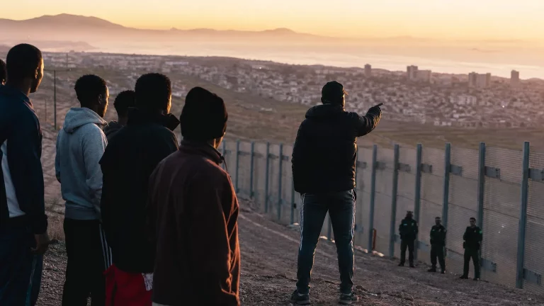Composición editorial sobria: siluetas de personas junto a una valla fronteriza, mientras una figura en perfil parcial, que sugiere una autoridad marroquí, parece observar. En el fondo, una bandera española ondea, y figuras difuminadas de la Guardia Civil se ven pasivas.