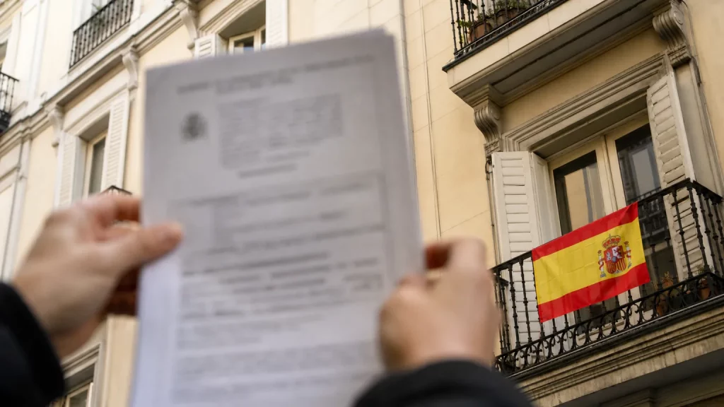 Bandera española ondeando en un balcón de un edificio residencial, con un documento oficial en primer plano.