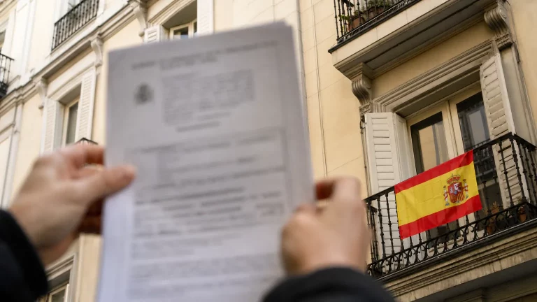 Bandera española ondeando en un balcón de un edificio residencial, con un documento oficial en primer plano.