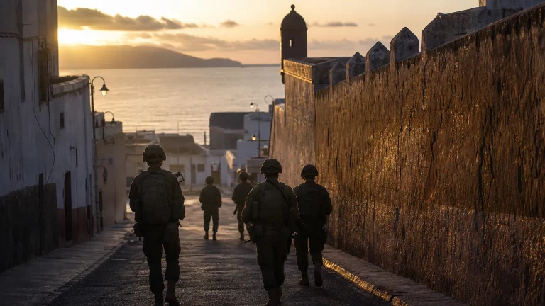 Siluetas de soldados con uniforme español en un puesto fronterizo, observando hacia el horizonte, con un paisaje urbano de fondo que sugiere Ceuta o Melilla