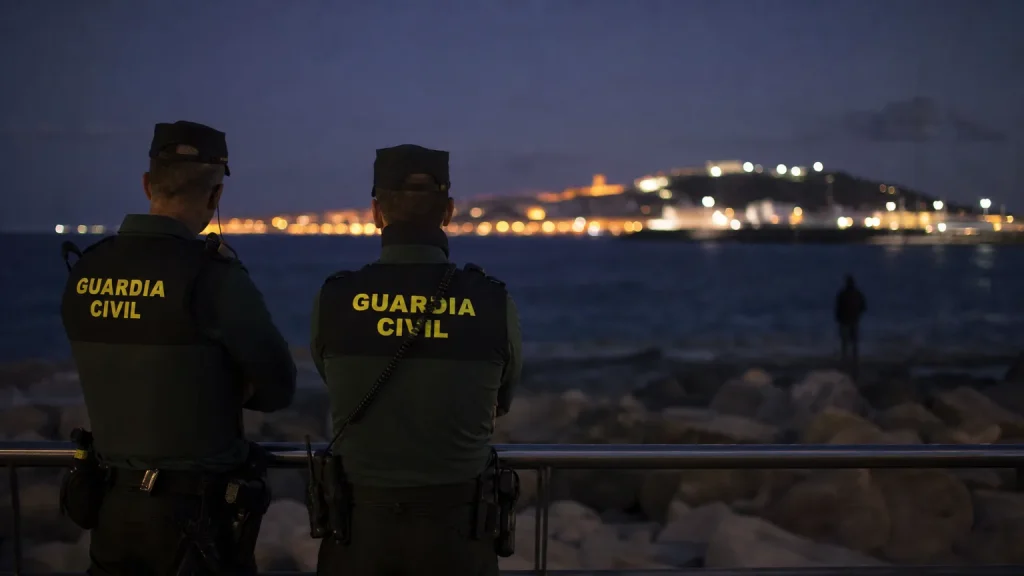 Agentes de la Guardia Civil o Policía Nacional de espaldas, vigilando una frontera marítima al anochecer en Ceuta, con luces difusas en el horizonte y una figura en la lejanía.