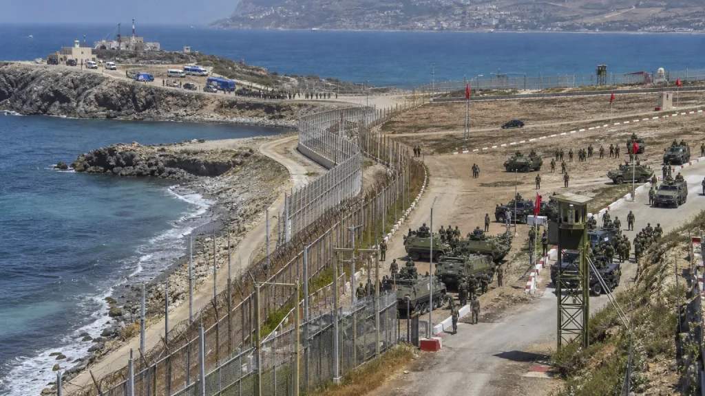 Soldados marroquíes y policía española en la frontera de Ceuta o Melilla, con armamento moderno al fondo y una valla fronteriza