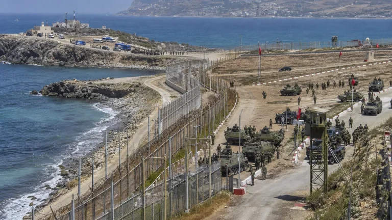 Soldados marroquíes y policía española en la frontera de Ceuta o Melilla, con armamento moderno al fondo y una valla fronteriza