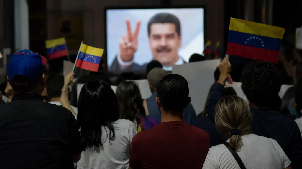 Siluetas de personas protestando frente a una pantalla donde se ve una imagen borrosa de Nicolás Maduro sonriendo y haciendo el signo de la victoria.