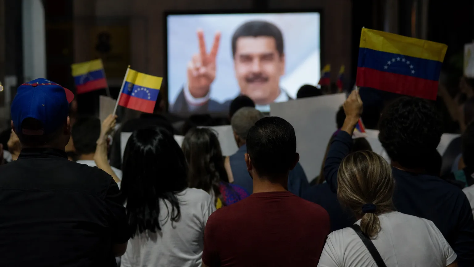 Siluetas de personas protestando frente a una pantalla donde se ve una imagen borrosa de Nicolás Maduro sonriendo y haciendo el signo de la victoria.
