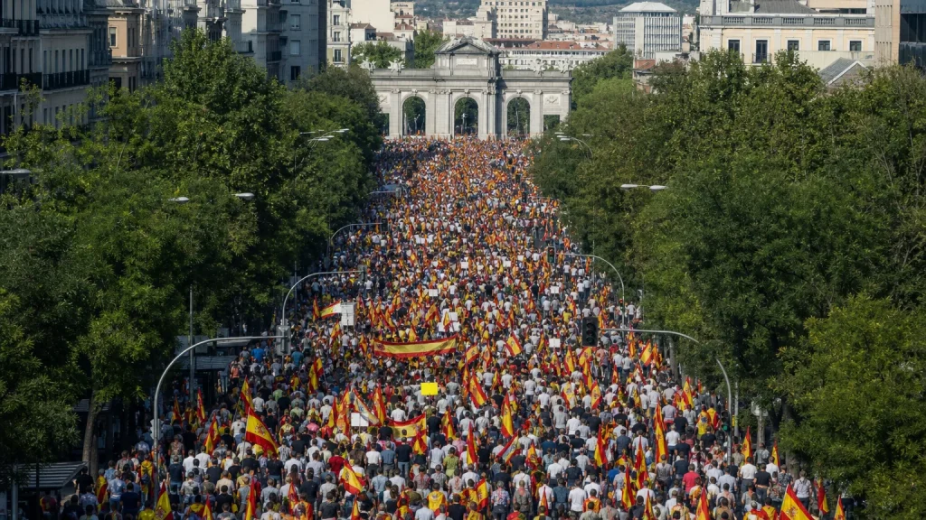 Vista aérea de una multitud marchando por una gran avenida de Madrid, con pancartas visibles pero ilegibles, hacia el Arco de la Victoria. Se aprecian figuras humanas de espaldas o en perfil, con banderas de España ondeando.