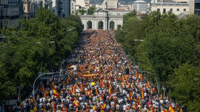 Vista aérea de una multitud marchando por una gran avenida de Madrid, con pancartas visibles pero ilegibles, hacia el Arco de la Victoria. Se aprecian figuras humanas de espaldas o en perfil, con banderas de España ondeando.