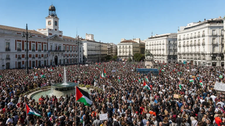 Vista aérea de una multitudinaria manifestación en una plaza céntrica de Madrid, con pancartas genéricas que expresan descontento ciudadano. Se aprecian edificios históricos al fondo.
