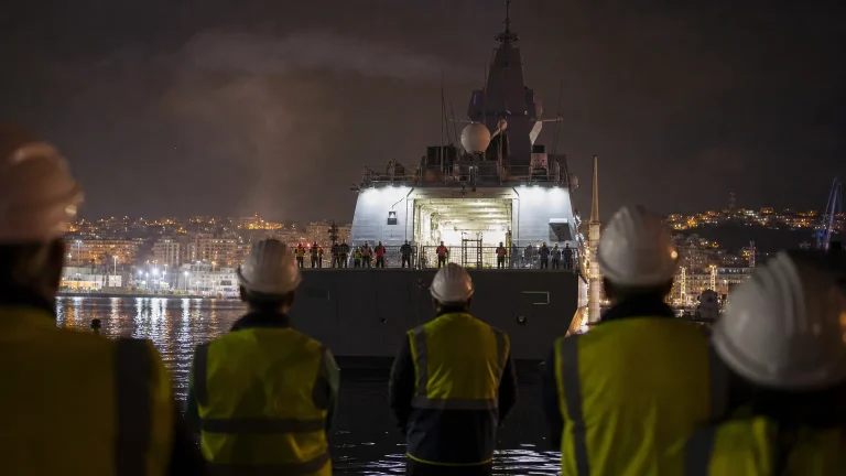 Silueta de un patrullero militar siendo botado en un astillero, con trabajadores de espaldas observando, y en el fondo, una bandera de Marruecos ondeando discretamente, simbolizando la entrega polémica