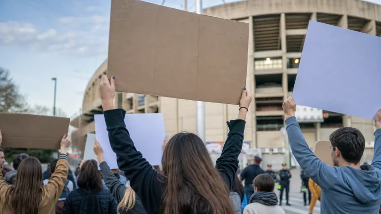 Personas sosteniendo pancartas en una manifestación pacífica frente a un estadio de fútbol, con algunas banderas de España visibles en segundo plano y los rostros desenfocados