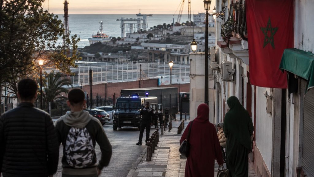 Escena editorial en una calle de Ceuta con tensión fronteriza y presencia marroquí simbólica